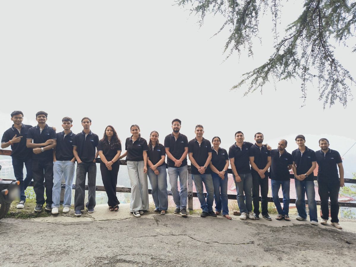 Fifteen members of the Netgen team in matching black shirts, standing in a row along a railing on a Shimla hillside under deodar branches