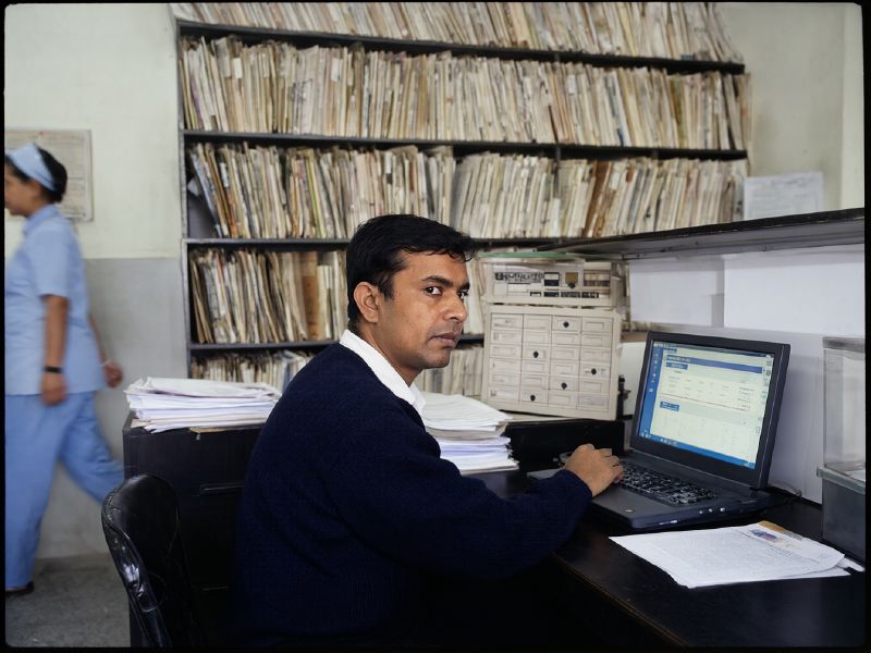 Photo (AI-generated, illustrative): a hospital records administrator working on a patient dashboard in front of shelves of paper files